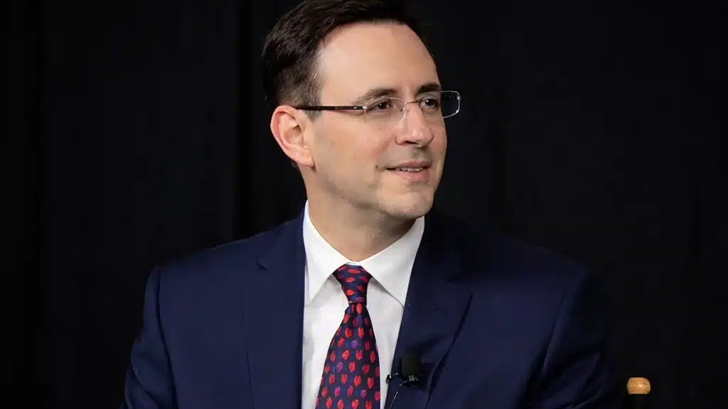 Yoram Eilon, seated, in a dark suit against a black backdrop